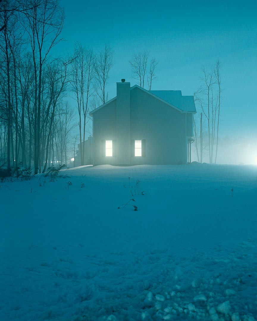 A suburban house at night in fog, a single window lit warm against cold blue-grey snow and bare trees.
